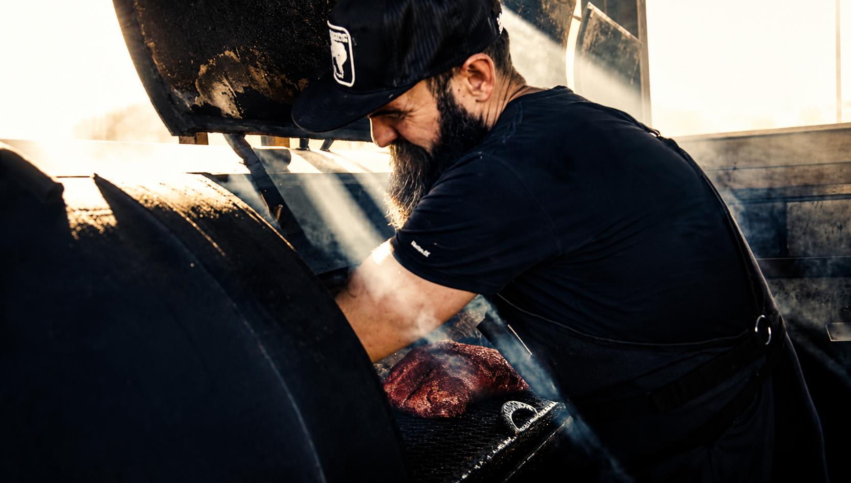 Colby Brownlee loading an offset smoker at sunrise, smoke drifting across the open cook chamber.