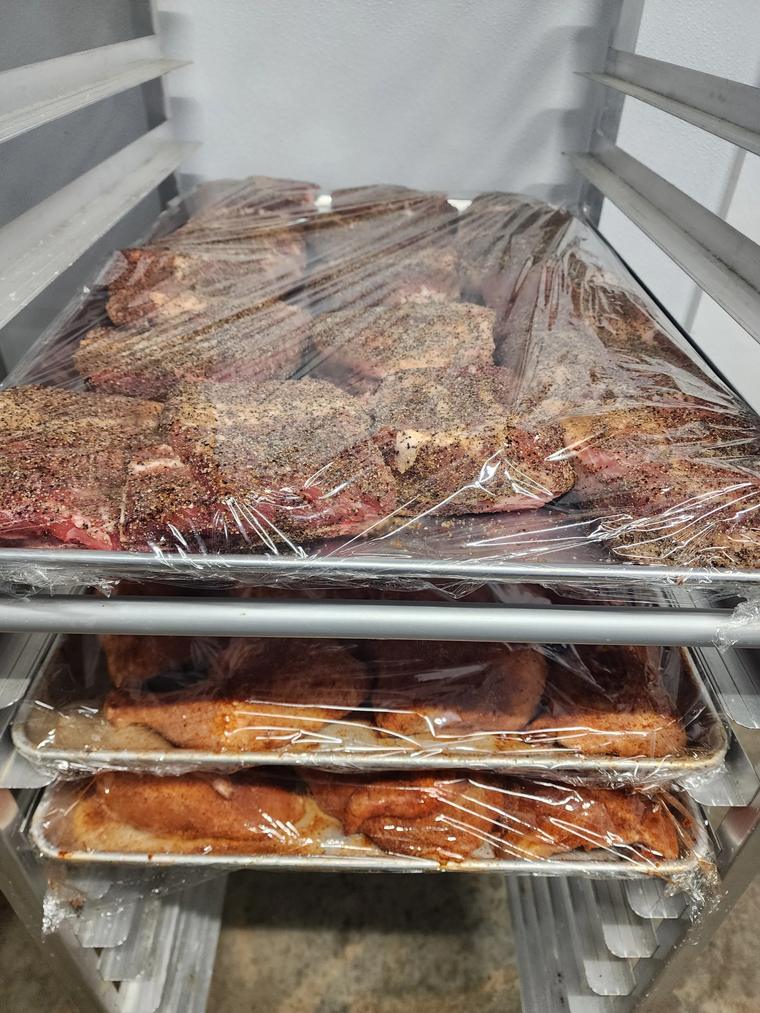 Three tiers of a speed rack holding wrapped sheet pans of seasoned protein, staged in cold storage.
