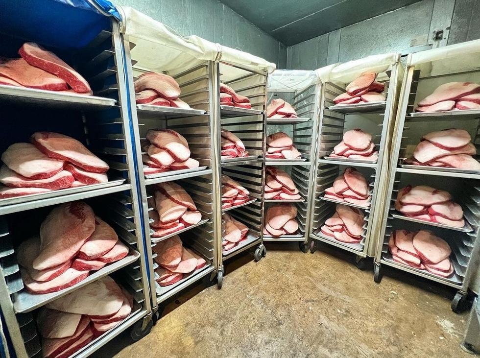 Production photograph from a separate location: briskets on sheet pans arranged on speed racks in a walk-in cooler.