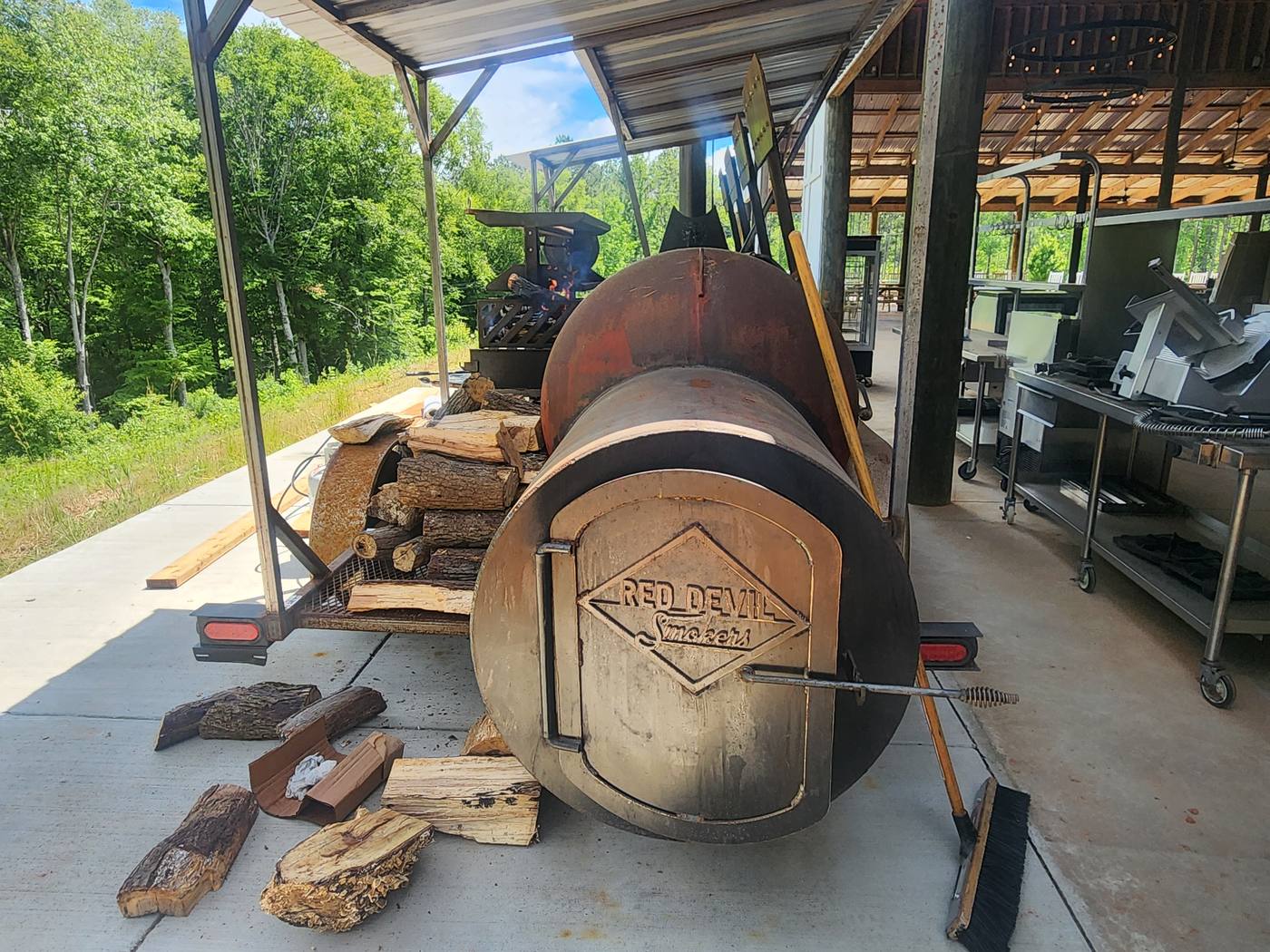Offset smoker on a trailer under a covered pavilion, split wood stacked alongside, kitchen equipment behind.