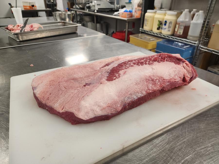 Untrimmed brisket packer on a white cutting board in a working kitchen, trim pan and knife behind.